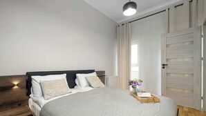A neatly made bed with a quilted cover, a bedside table with a lamp, and a window with curtains. - A Comfortable Apartment in the Centre of Poznań | Close to the MTP (Poznań)