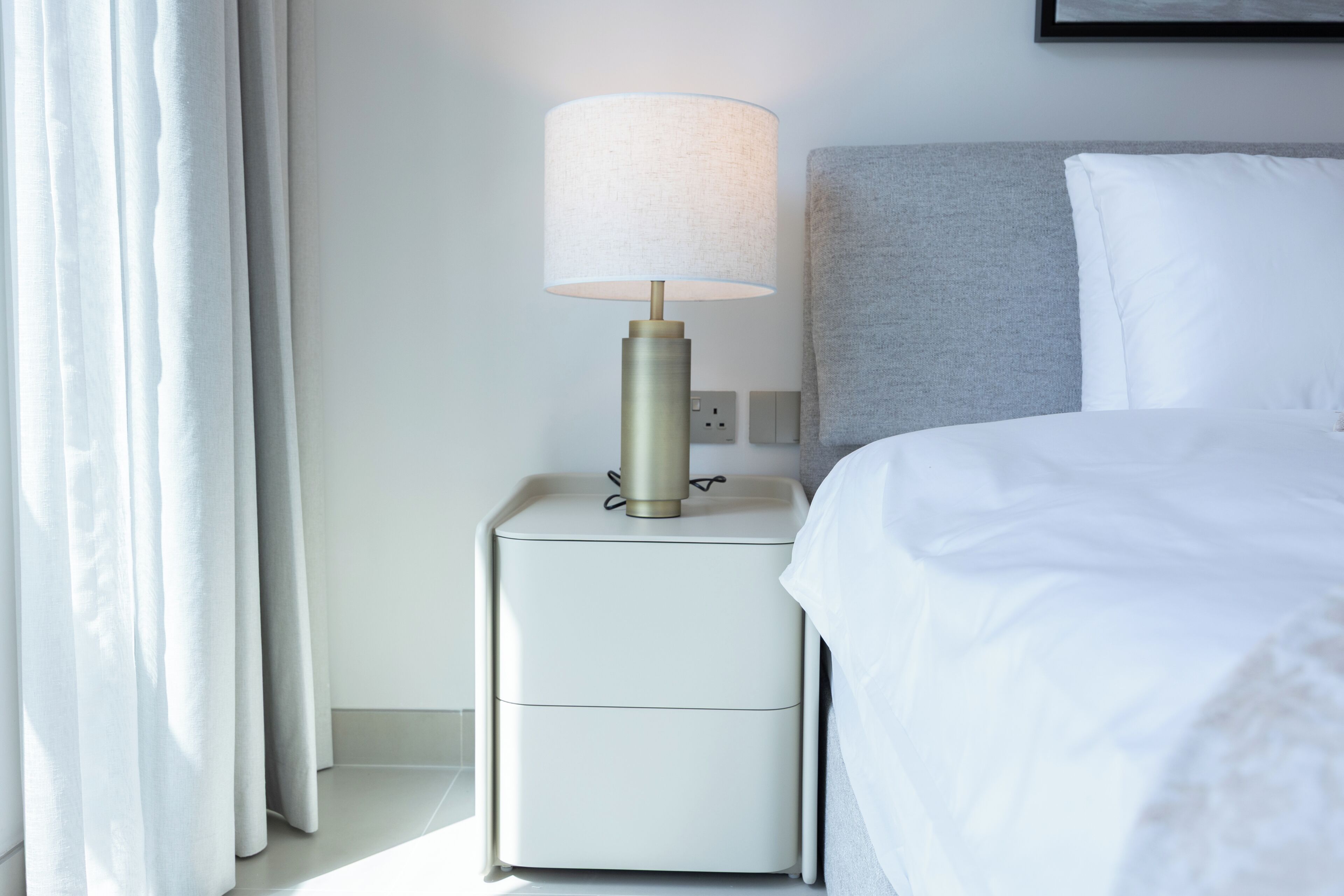 Ein ordentlich gemachtes Bett mit weißer Bettwäsche, eine Nachttischlampe mit zylindrischem Lampenschirm und ein grauer Kopfteil.