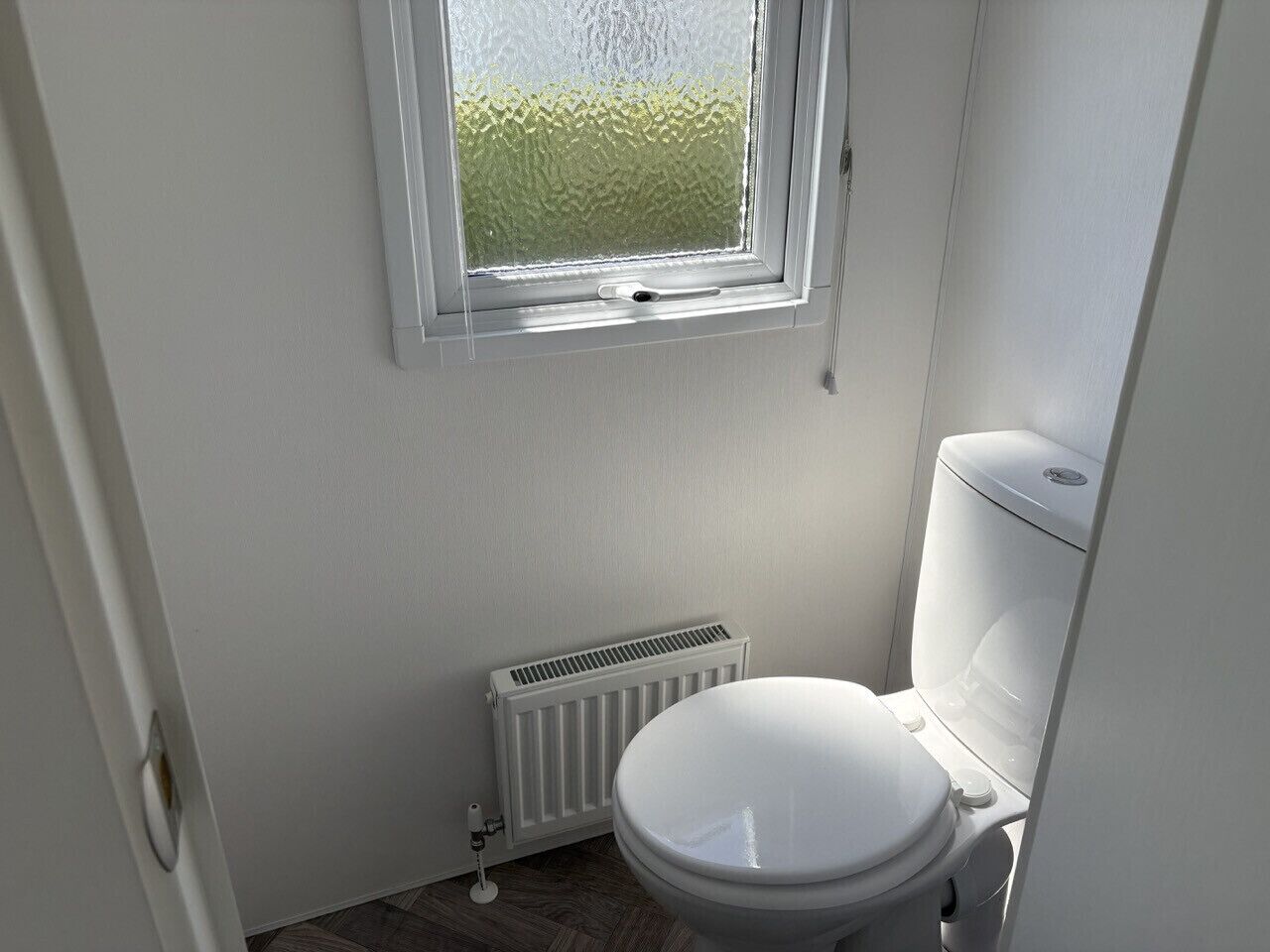 Un baño pequeño y limpio con inodoro blanco, un radiador y una ventana que da a zonas verdes.