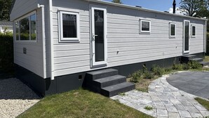 A single-story mobile home with a front porch, a small front yard, and a paved driveway. - De Zeeuwse Parel Vacation Park - Sunset Comfort (Scherpenisse)