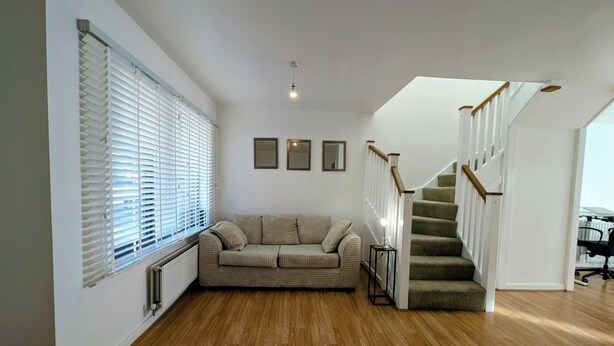 A spacious living area with a wooden floor, a beige sofa, a staircase with a wooden handrail, and a radiator near the window. - Modern 2BDR Garden Home With Parking and WiFi (Greater London)
