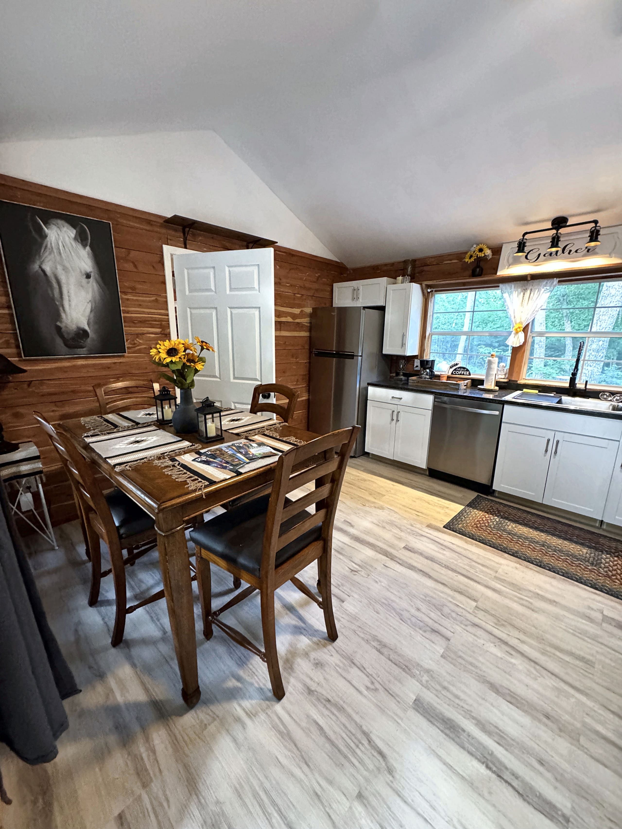 A dining area with a wooden table and chairs, a kitchen with white cabinets, a stainless steel refrigerator, and a large window with a view of greenery.