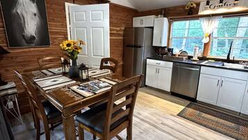 A dining area with a wooden table and chairs, a kitchen with white cabinets, a stainless steel refrigerator, and a large window with a view of greenery.