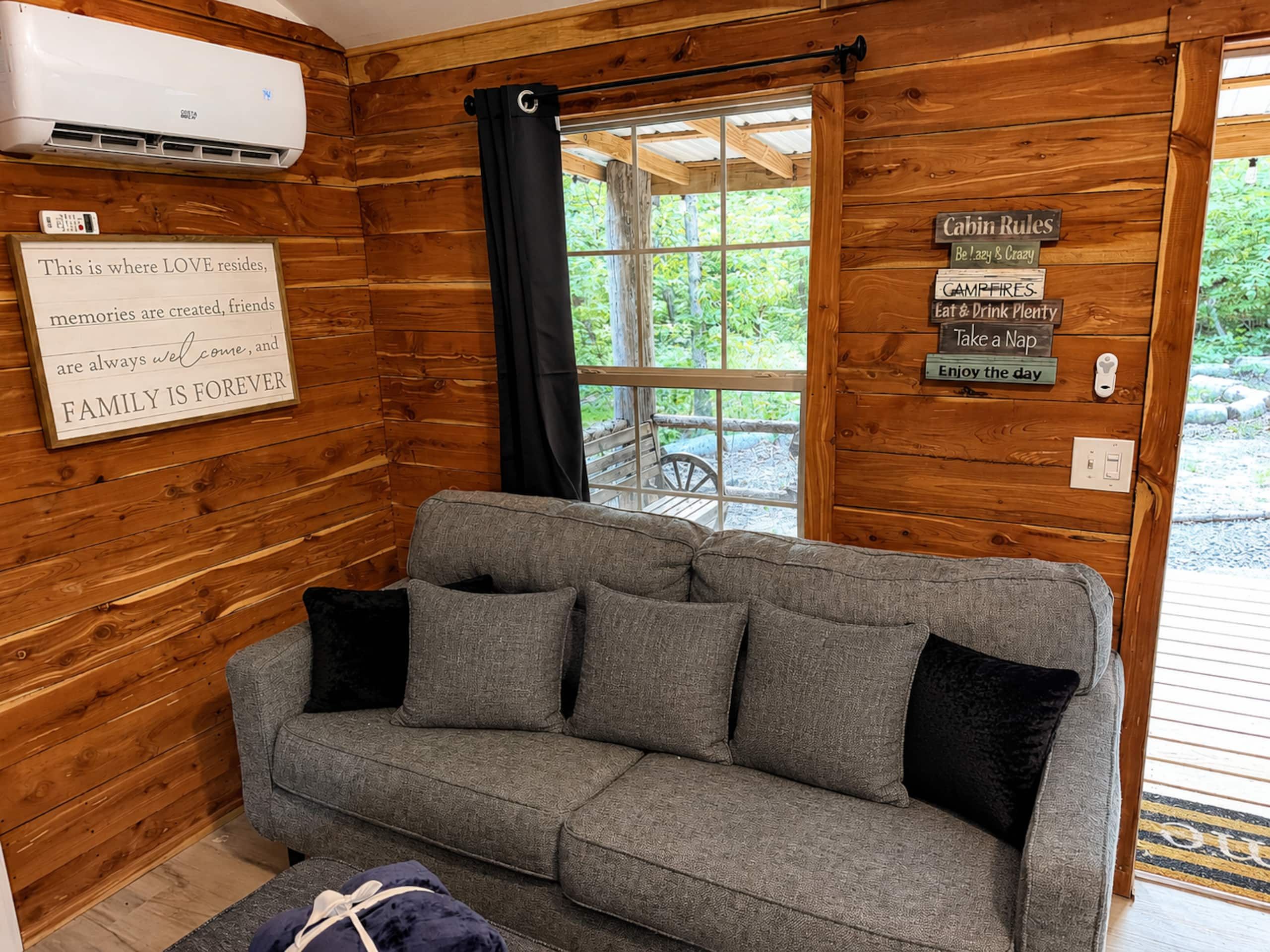 Interior acollidor d'una cabana amb un sofà gris, un cartell que diu 'Aquest és on resideix l'AMOR' i una finestra amb vista a verdura.
