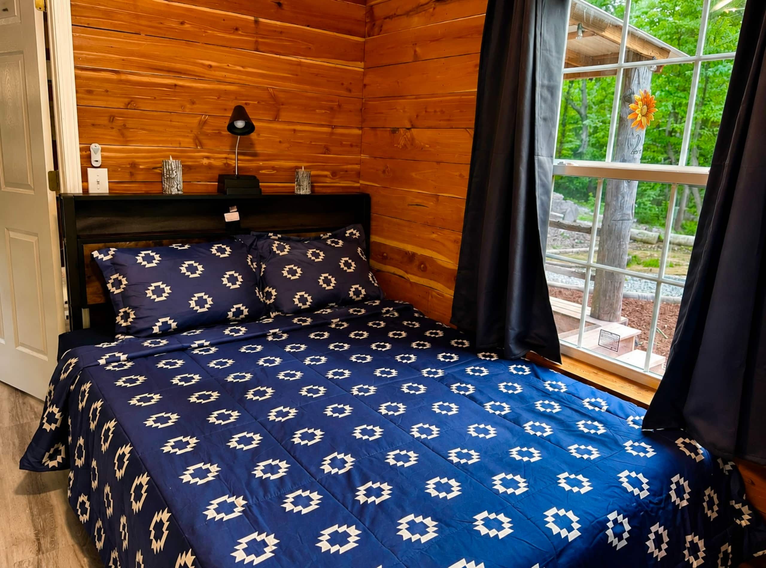 Habitació de cabana de fusta amb un llit, una tauleta de nit, una làmpada i una finestra amb vista a vegetació.