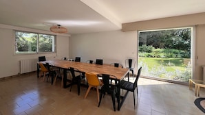 Un espace repas comprenant une longue table, des chaises et une vue sur un jardin, grâce à de grandes fenêtres.