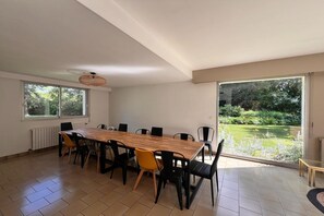 A dining area with a long table, chairs, and a view of a garden through large windows. - Grand French Family Holiday Home (Saint-Germain sur Bresle)