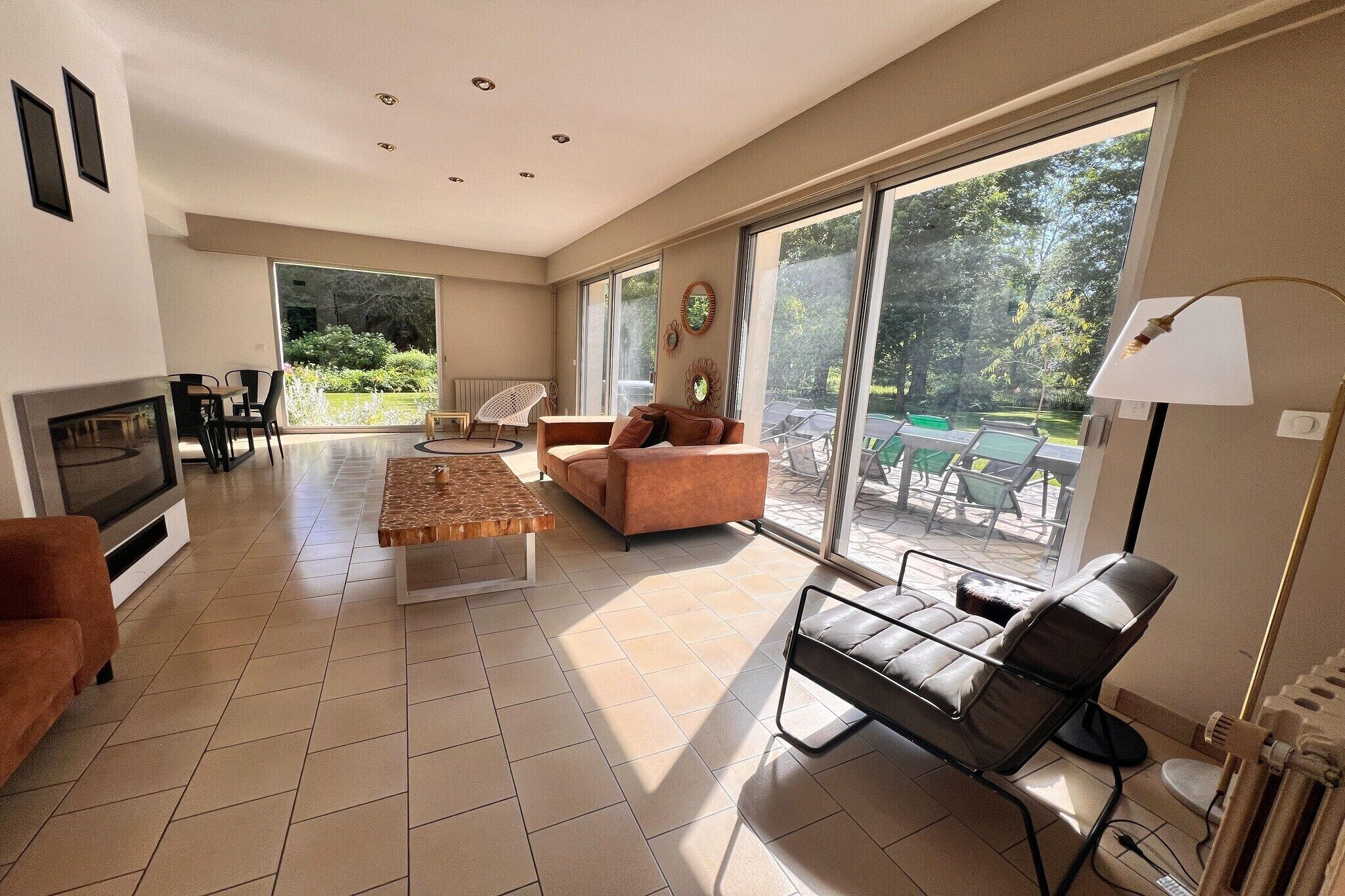 A spacious living room with a fireplace, a sofa, a coffee table, and a TV. There are large windows providing a view of the outdoors.