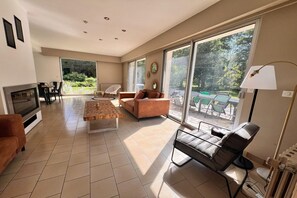 A spacious living room with a fireplace, a sofa, a coffee table, and a TV. There are large windows providing a view of the outdoors. - Grand French Family Holiday Home (Saint-Germain sur Bresle)