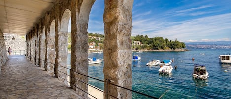A stone-paved walkway with arches, a clear blue sky, and boats on a calm sea.