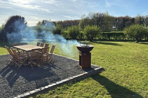 An outdoor dining area with a table and chairs, surrounded by a gravel border, and a barbecue grill emitting smoke. - Hoefke Voort – Rural farmhouse in Haspengouw, total peace in protected landscape (Tongeren-Borgloon)