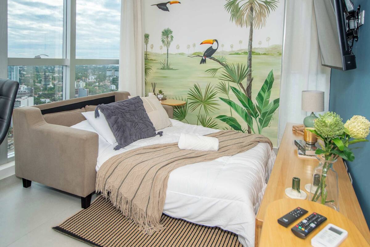 Ein Hotelzimmer mit Bett, Sofa, Schreibtisch mit Lampe, Blumen in einer Vase und Blick auf die Stadt durch große Fenster.