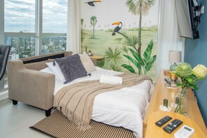 Ein Hotelzimmer mit Bett, Sofa, Schreibtisch mit Lampe, Blumen in einer Vase und Blick auf die Stadt durch große Fenster.