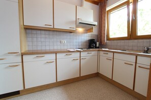 Ett kök med vita skåp, trähandtag, en kakelbeklädd vägg bakom arbetsbänken, ett fönster med gardiner och en kaffebryggare på bänken.