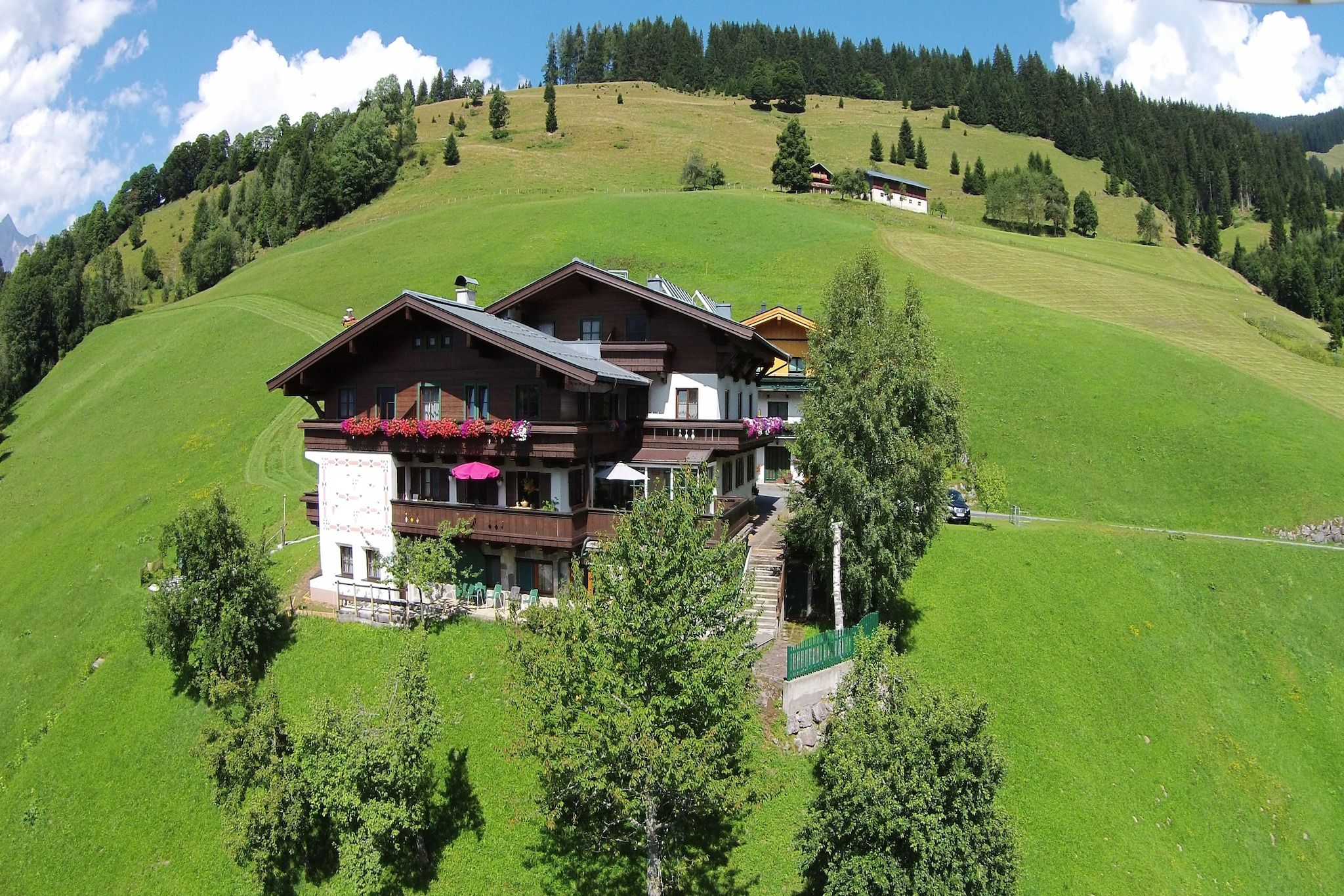Un chalet à deux étages doté d’un balcon, entouré de collines verdoyantes et d’arbres luxuriants.
