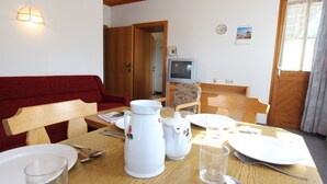 Un'area pranzo con un tavolo in legno apparecchiato per due persone, un divano rosso, una televisione e una porta che dà su un'altra stanza.