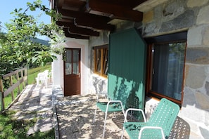 Un espace extérieur abrité doté d’une terrasse en bois, d’un mur en pierre et d’une porte donnant sur une pièce. On y trouve deux chaises vertes et une table blanche.