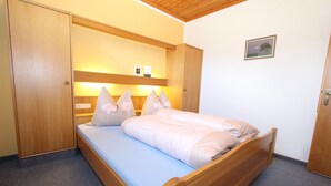 Un letto singolo con testiera e pediera in legno, un materasso azzurro chiaro e diversi cuscini. A sinistra un armadio in legno e a destra una porta.