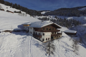 Un chalet enneigé à la façade en bois, entouré d’un paysage hivernal et de collines boisées.