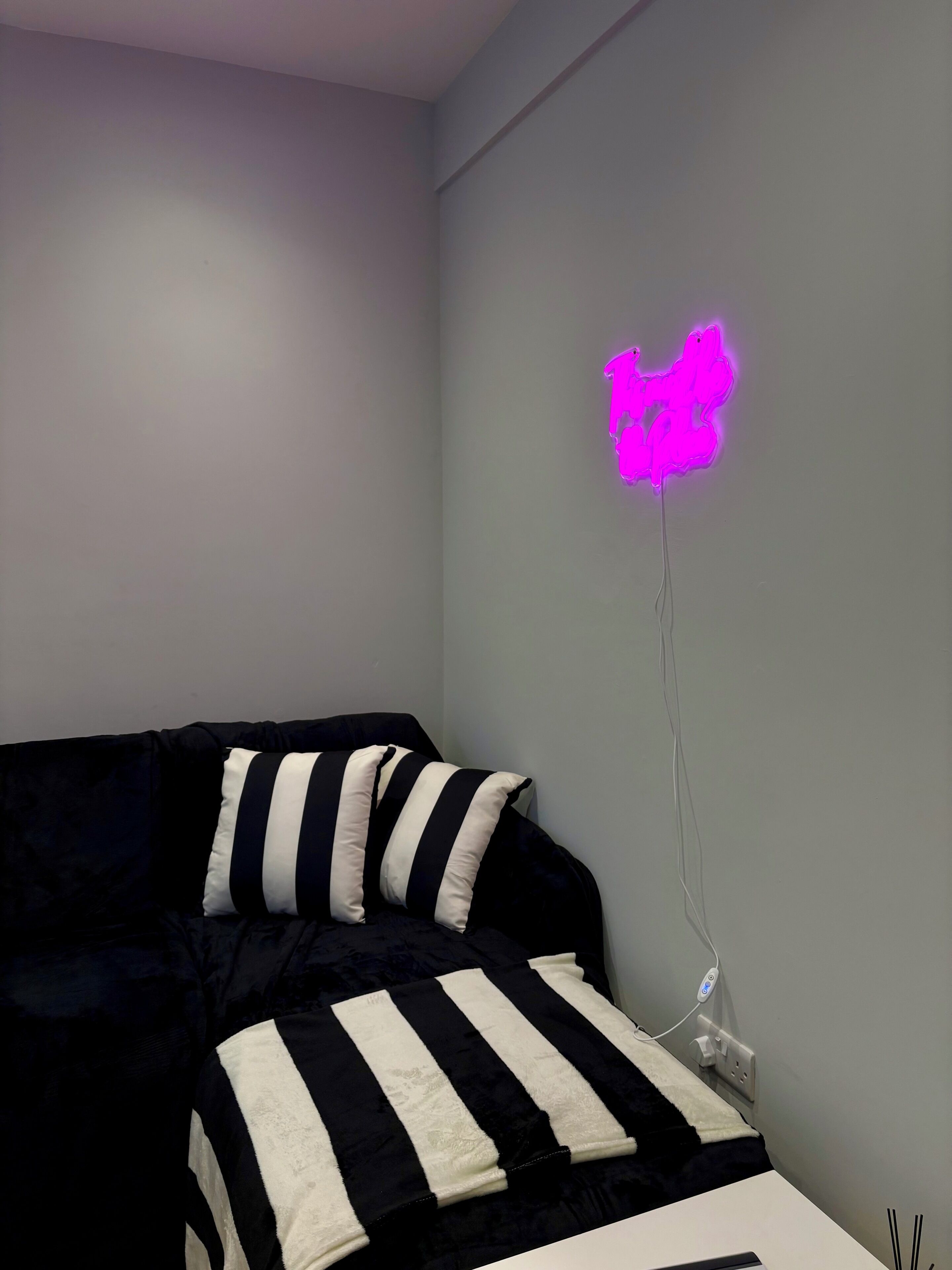 Et soveværelse med en seng med stribet sengetøj i sort og hvid, en lysarmatur i neonrosa på væggen, og en stikkontakt.