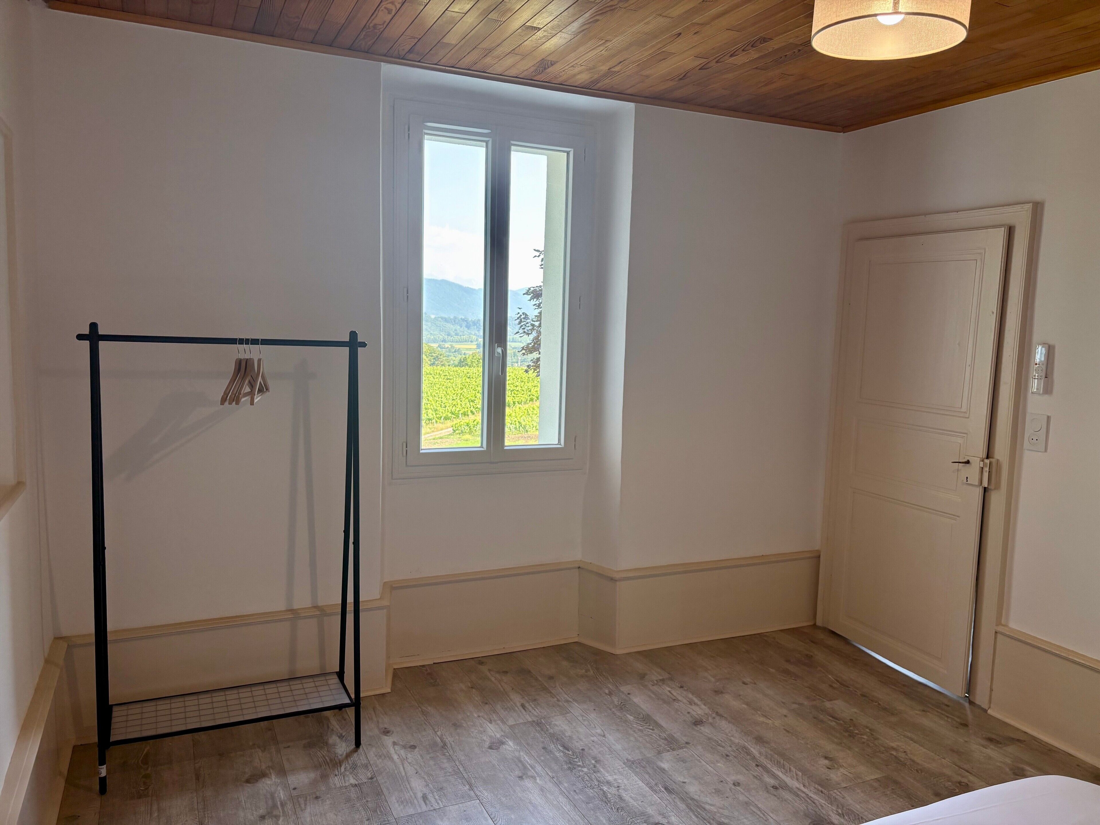 Habitación con piso de madera, ventana con vista a follaje y una puerta, además de un estante de metal.