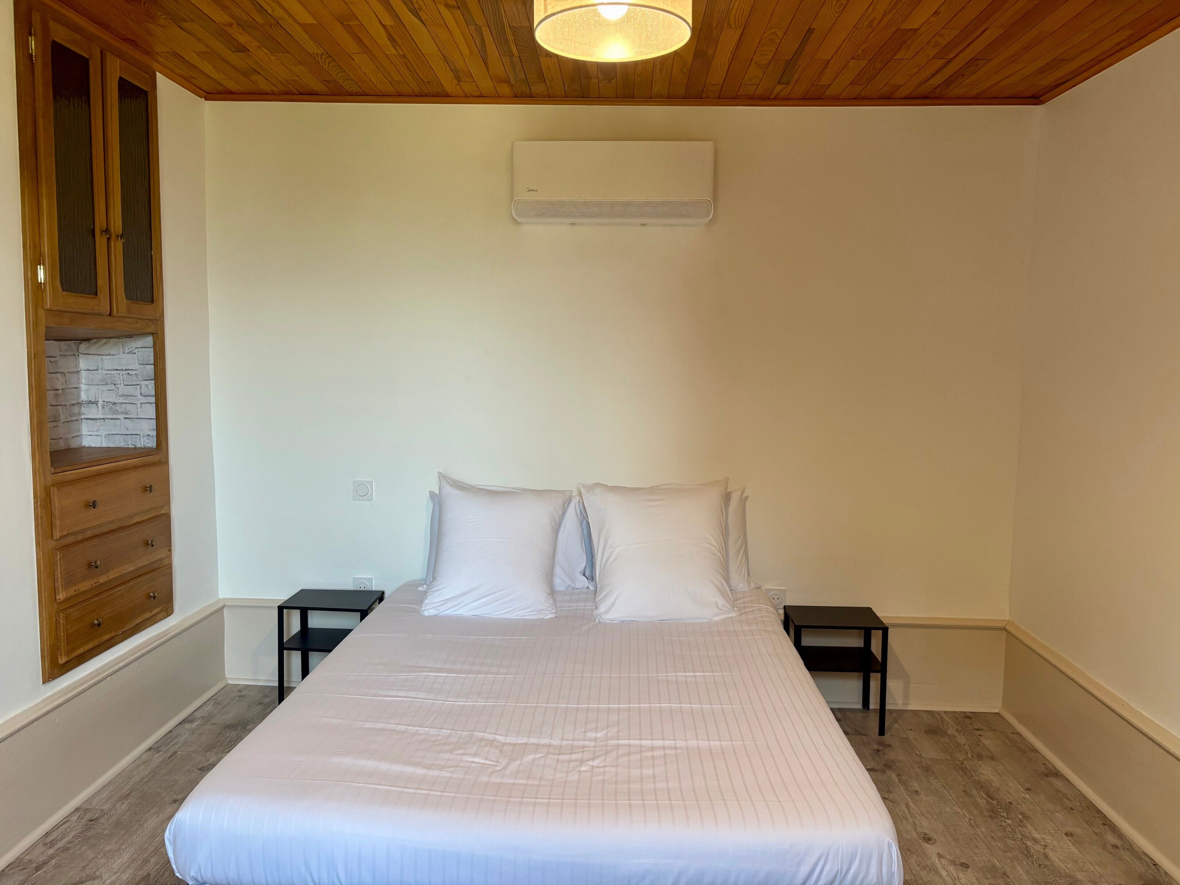 Une chambre à coucher comprenant un lit, une armoire en bois, des tables de chevet et un appareil de climatisation.