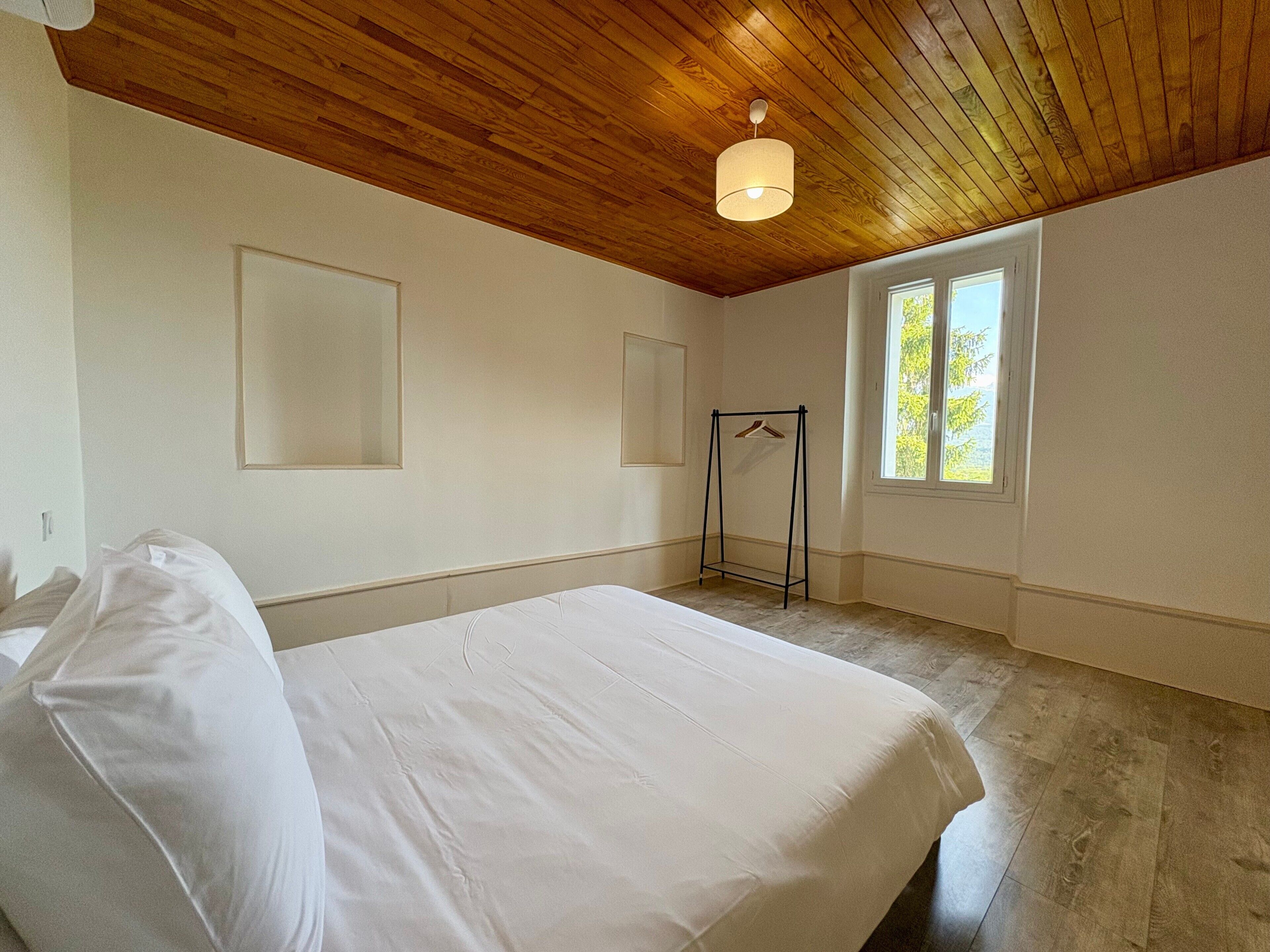A bedroom with a wooden ceiling, a bed with white linens, a window, and a wall-mounted coat rack.