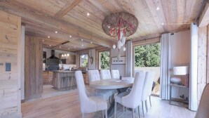 Ein modernes Interieur mit Holzbalkendecke, einem runden Esstisch mit Stühlen, einer Küchenzeile und Blick durch große Fenster.