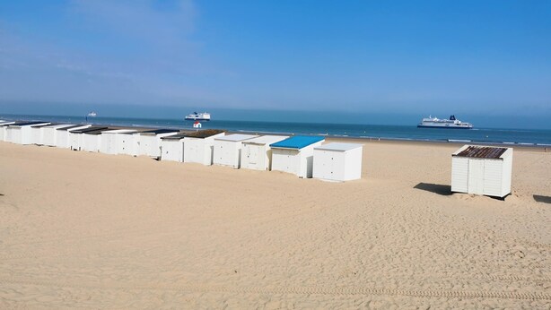 Ein Strand mit mehreren weißen Strandhütten, die in einer Reihe aufgereiht sind, ein großes Schiff in der Ferne und ein strahlend blauer Himmel.