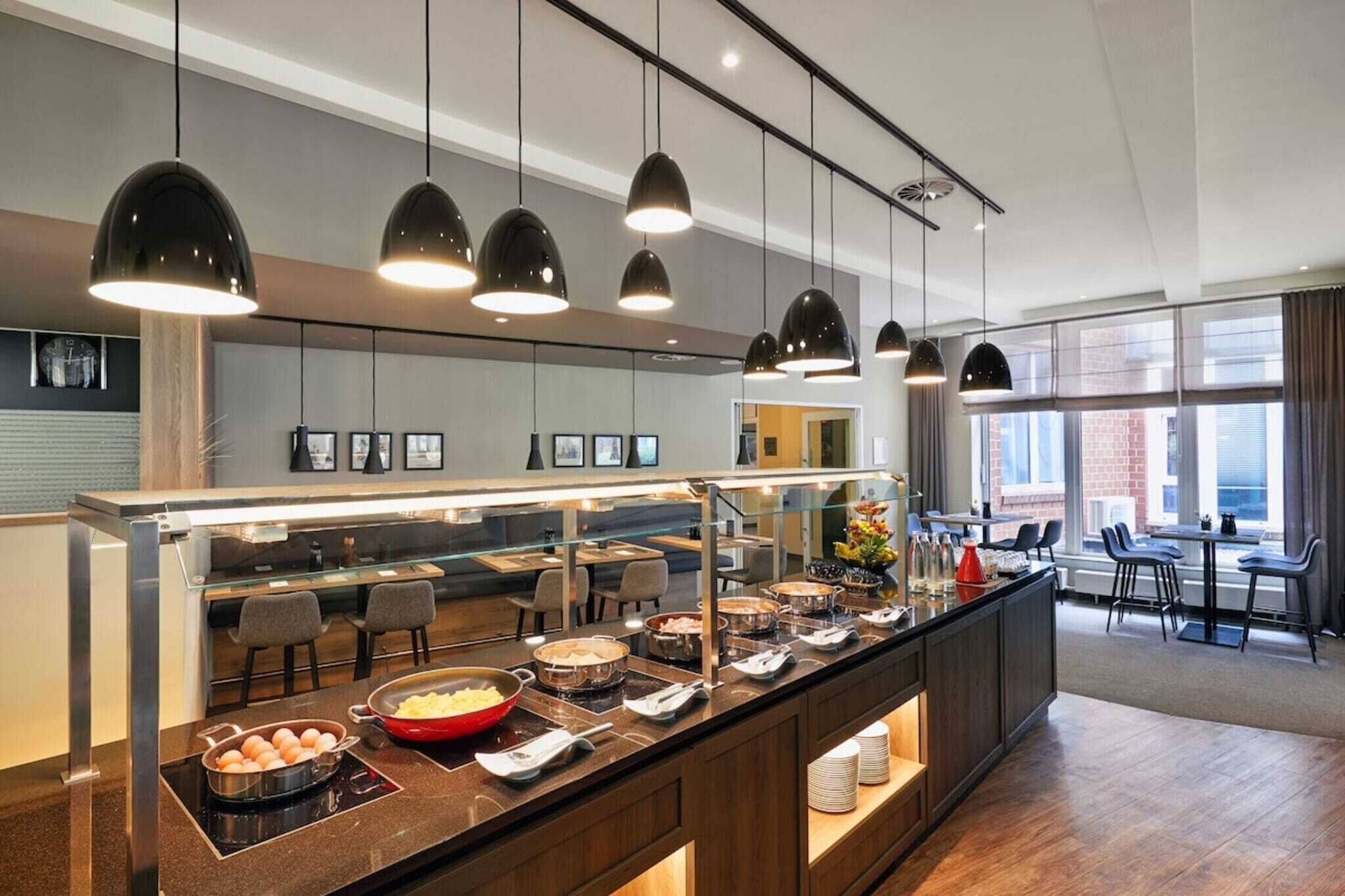 A modern hotel breakfast area with a buffet, pendant lighting, and seating.