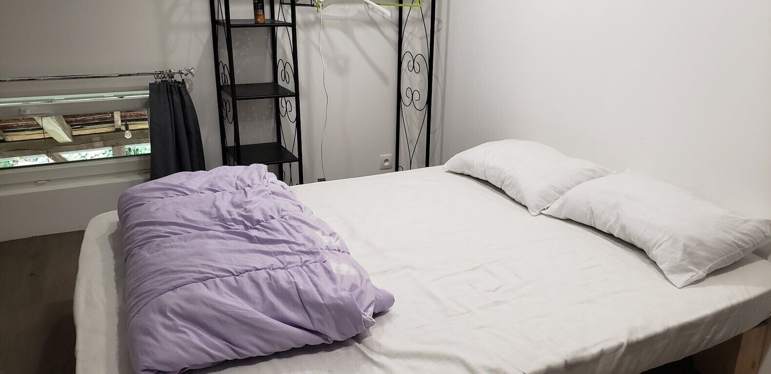 Een bed met witte lakens, een paars dekbed en een metalen plankunit in zwart, tegen een witte muur.