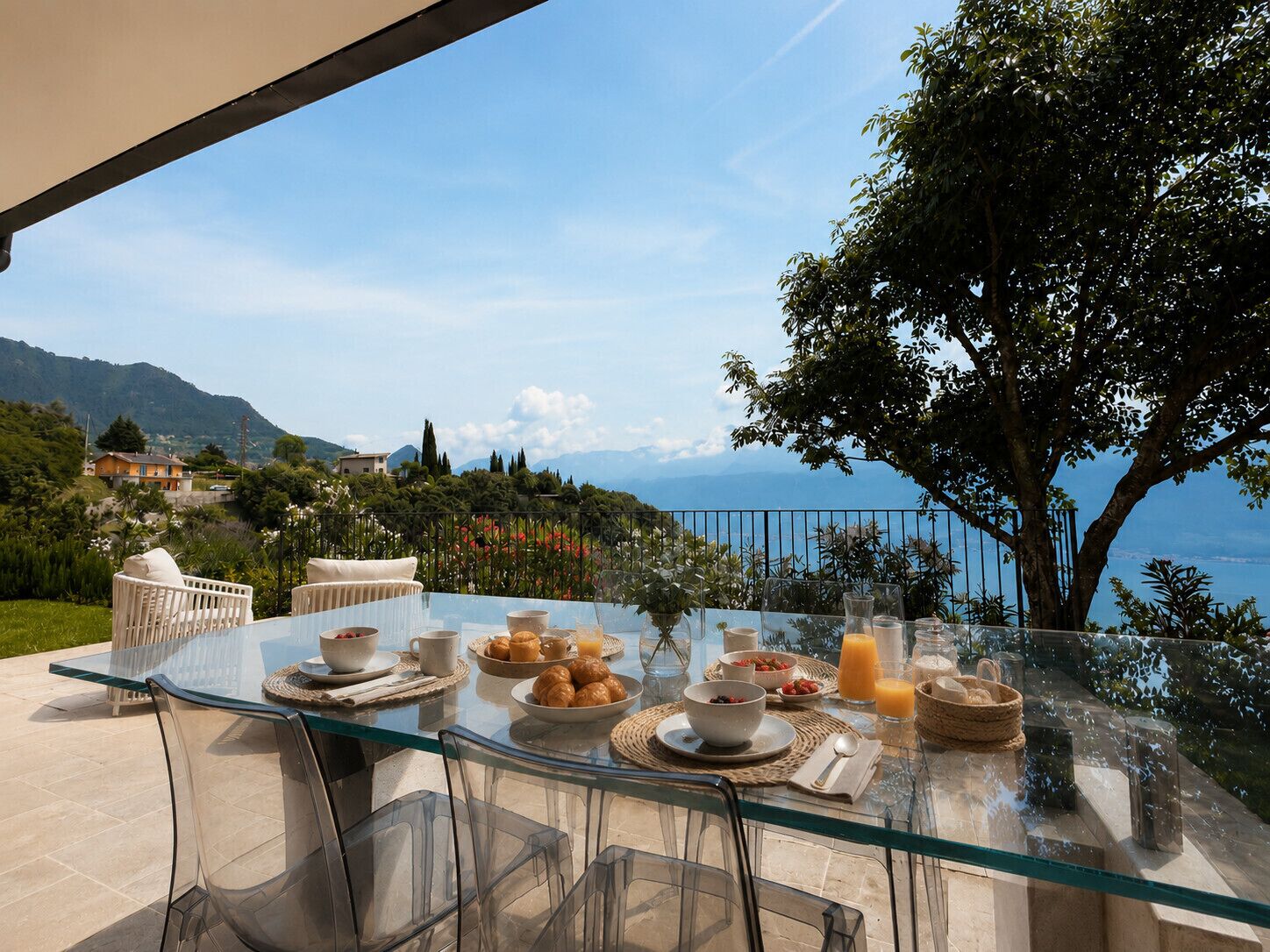 A glass table with breakfast items, clear chairs, and a scenic view of the sea and mountains.