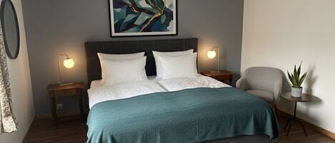 A modern bedroom with a bed, bedside lamps, a gray headboard, a framed abstract painting, a gray armchair, and a small potted plant.