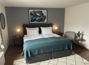 A modern bedroom with a bed, bedside lamps, a gray headboard, a framed abstract painting, a gray armchair, and a small potted plant. - Les Vieux Toits (Laténa)