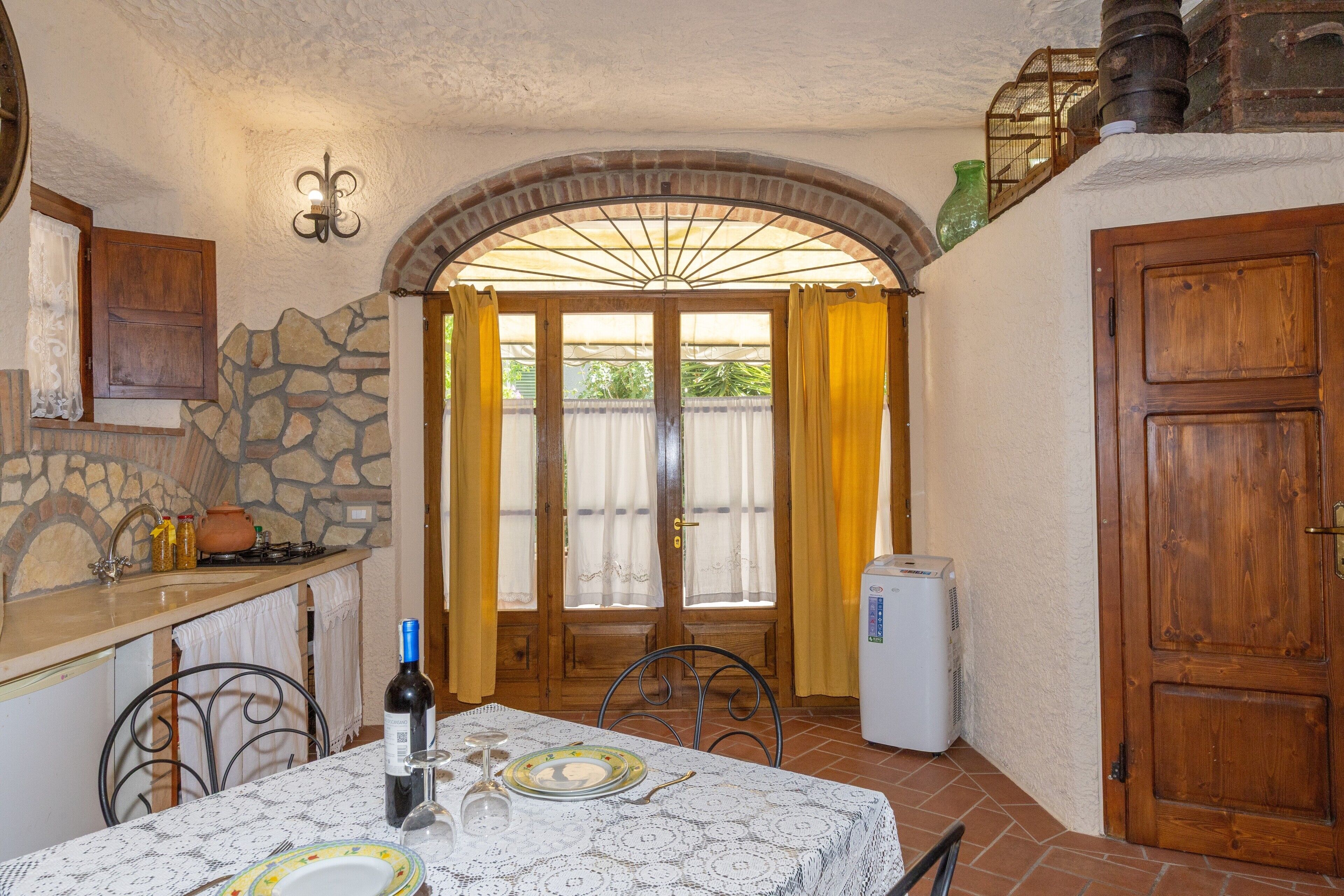 A rustic kitchen with a stone wall, wooden cabinets, a table set for dining, and a view through a glass door.