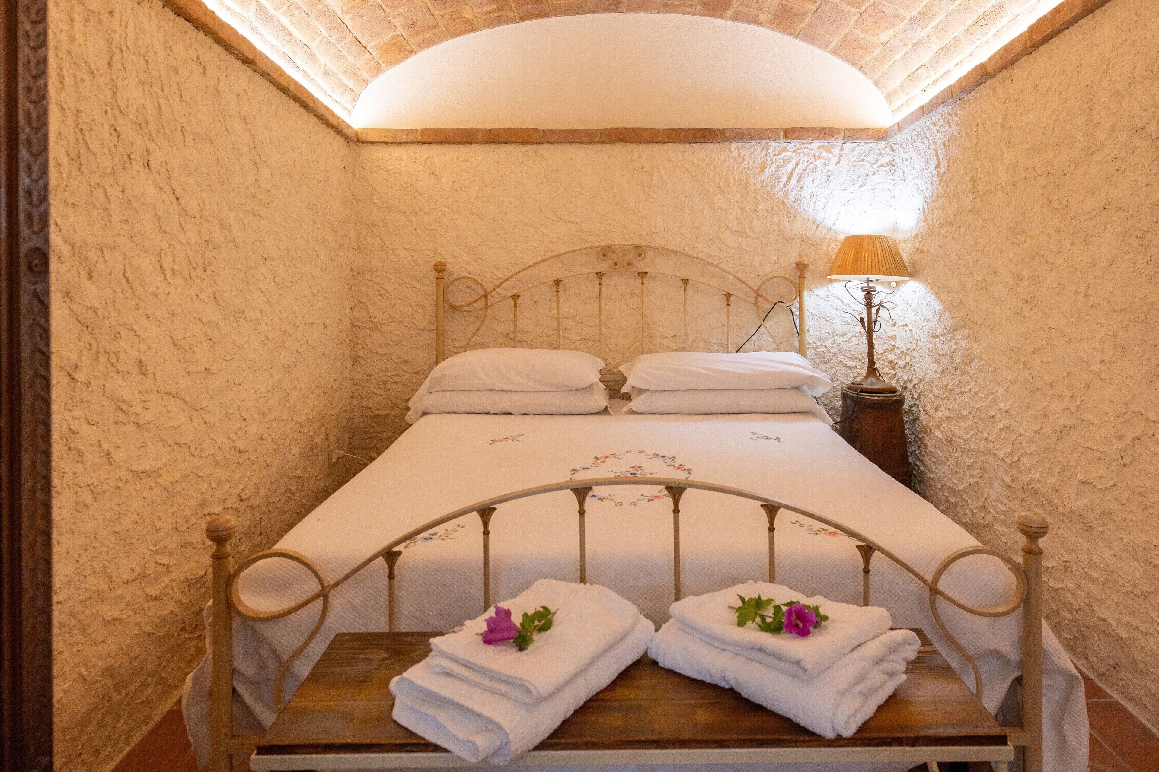 A single bed with a headboard, a bedside table with a lamp, and two towels with flowers on a wooden bench.