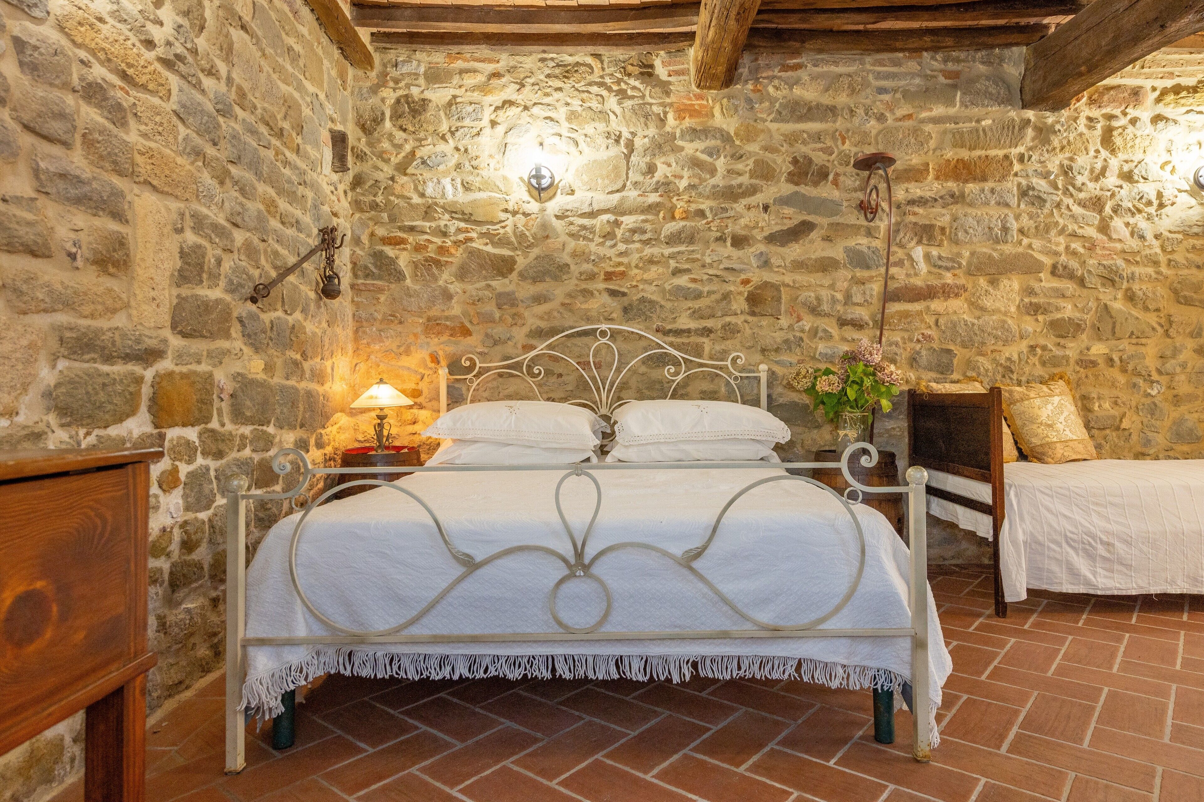 A stone-walled bedroom with a bed, bedside table, lamp, and a small plant.