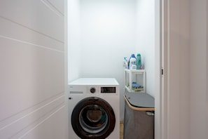 Une petite buanderie équipée d’un lave-linge à chargement frontal, d’une petite étagère contenant des produits de nettoyage et d’un banc gris.