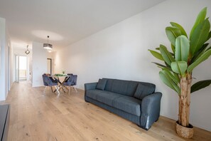 Ein modernes Wohnzimmer mit einem grauen Sofa, einem Esstisch mit Stühlen, einer großen Topfpflanze und einer Pendelleuchte.