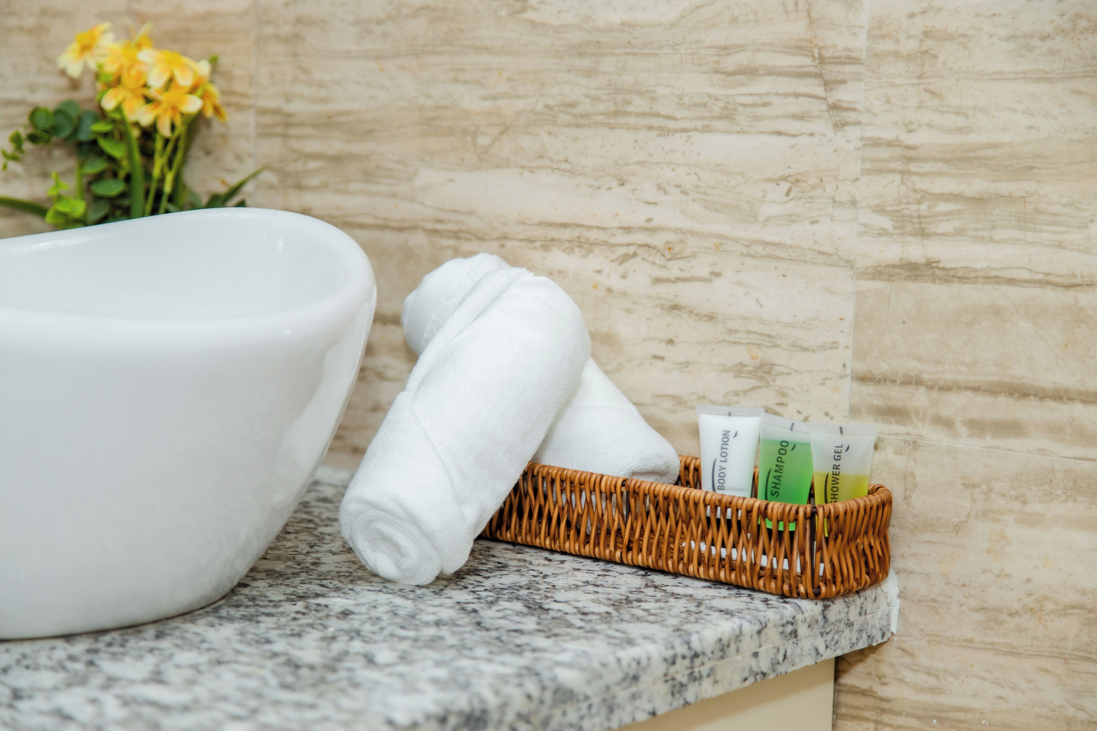 A bathroom counter with a white basin, a rolled-up towel, and a basket containing toiletries.
