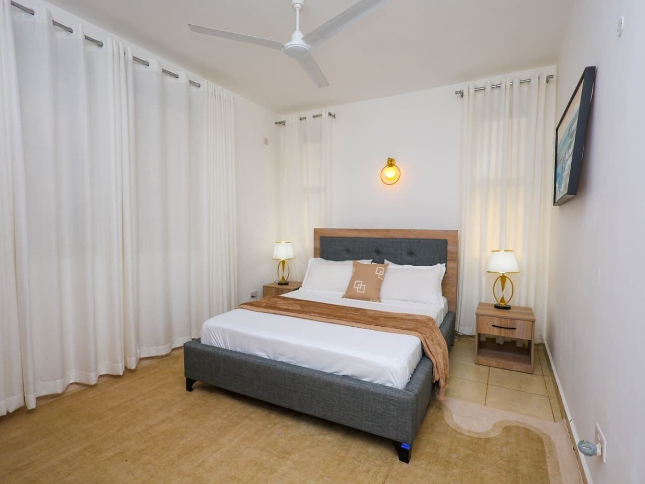 A hotel room with a bed, two bedside tables, a ceiling fan, and a wall-mounted light fixture.