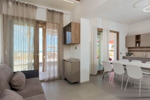 Un salon moderne comprenant un canapé, une table à manger avec des chaises, un coin cuisine avec des placards et une vue sur l’extérieur grâce à une porte vitrée.