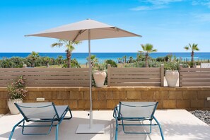 A patio with two chairs and a large umbrella, overlooking a beach with palm trees and a clear blue sky. - Wonderful seafront villa with terrace in Marzamemi (Noto)