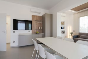 A modern kitchen with a built-in TV, white cabinets, and a dining area with a long table and chairs. - Wonderful seafront villa with terrace in Marzamemi (Noto)