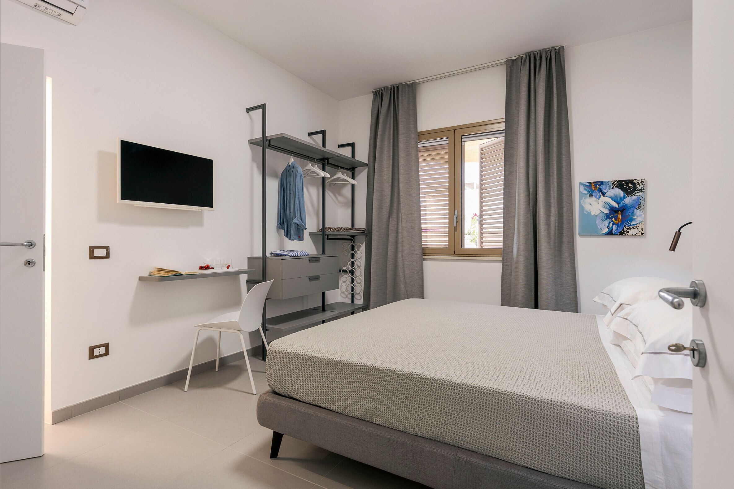 A bedroom with a bed, a desk, a chair, a TV, a window with curtains, and a painting on the wall.