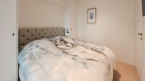 Un dormitorio con cabecera capitonada, cama con manta estampada, un cuadro en la pared y una puerta a la derecha.