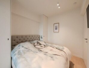 Ein Schlafzimmer mit einer gepolsterten Kopfteil, einem Bett mit gemustertem Überwurf, einem gerahmten Bild an der Wand und einer Tür rechts.