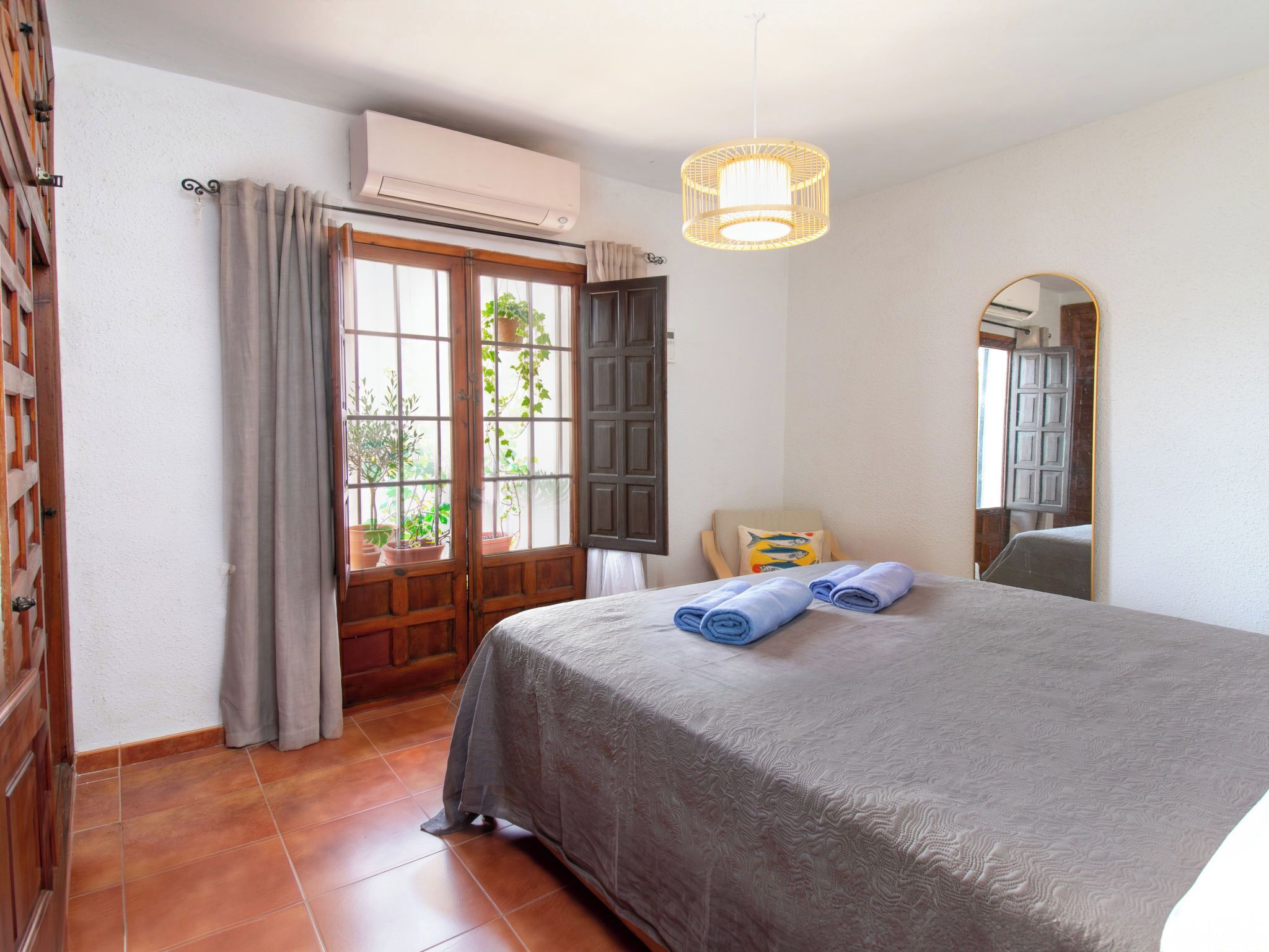 Une chambre à coucher avec un lit, une fenêtre avec des rideaux, un appareil de climatisation, un luminaire suspendu et un miroir.