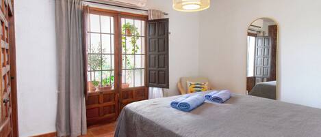 A bedroom with a bed, a window with curtains, an air conditioning unit, a hanging light fixture, and a mirror.