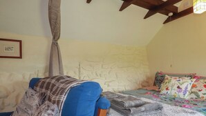 Quarto com parede de pedra, cama com edredom floral, cadeira azul com manta e vigas de madeira no teto.